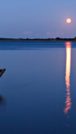 Singular chair in calm water facing the land in the horizon. With rising orange moon reflected in the blue water